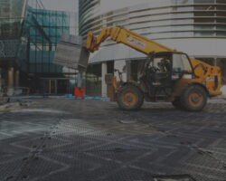 A Construction Site With A Yellow Forklift Moving A Large Metal Crate, Possibly Containing Products. The Site Is Surrounded By Modern Glass Buildings. Several Workers Are Visible In The Background, And The Ground Is Covered With Interlocking Metal Plates.
