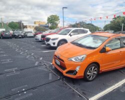An Outdoor Parking Lot With Various Cars Parked, Including An Orange And White Compact Car In The Foreground. The Ground Surface, Covered By ProtectaMats, Appears Metallic, And Red Triangular Flags Are Strung Overhead. The Background Shows Other Parked Cars And Buildings Under A Cloudy Sky.