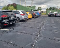 A Parking Lot With Multiple Cars Parked In A Row, Including A Black SUV, A Silver Crossover, And A Bright Yellow Car. The Ground Is Covered With ProtectaMats Modular Flooring Featuring Standard Tread. Green Industrial Buildings And Tall Chimneys Are Visible In The Background.